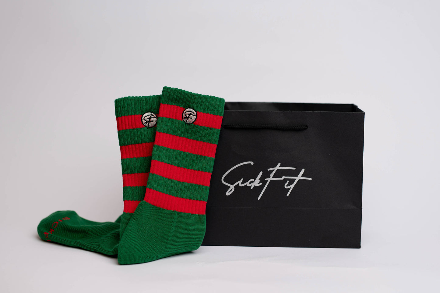 A pair of crew compression holiday socks with bold green and vibrant red stripes, placed on a white surface next to a black SickFit branded bag.