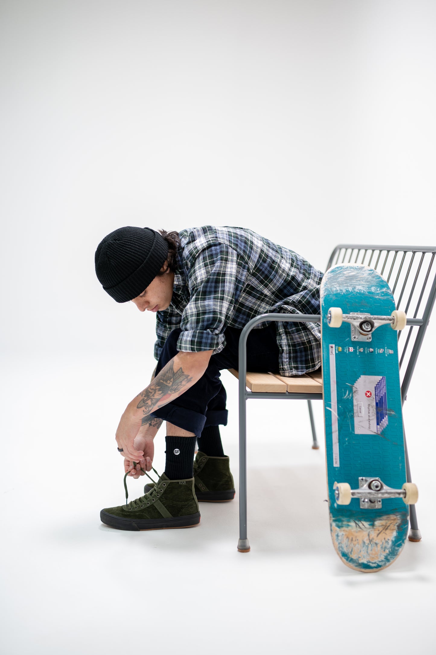 Person adjusting a shoe next to a blue skateboard on a white background wearing black socks.