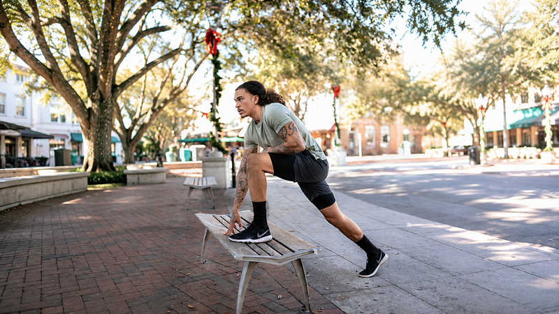 A man in athletic wear stretches on a bench in an outdoor urban plaza with trees and buildings in the background.