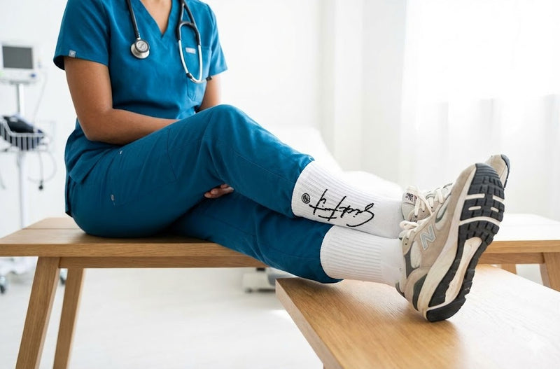 Person wearing blue scrubs sitting on a bench with feet up, wearing white socks and black and white sneakers.